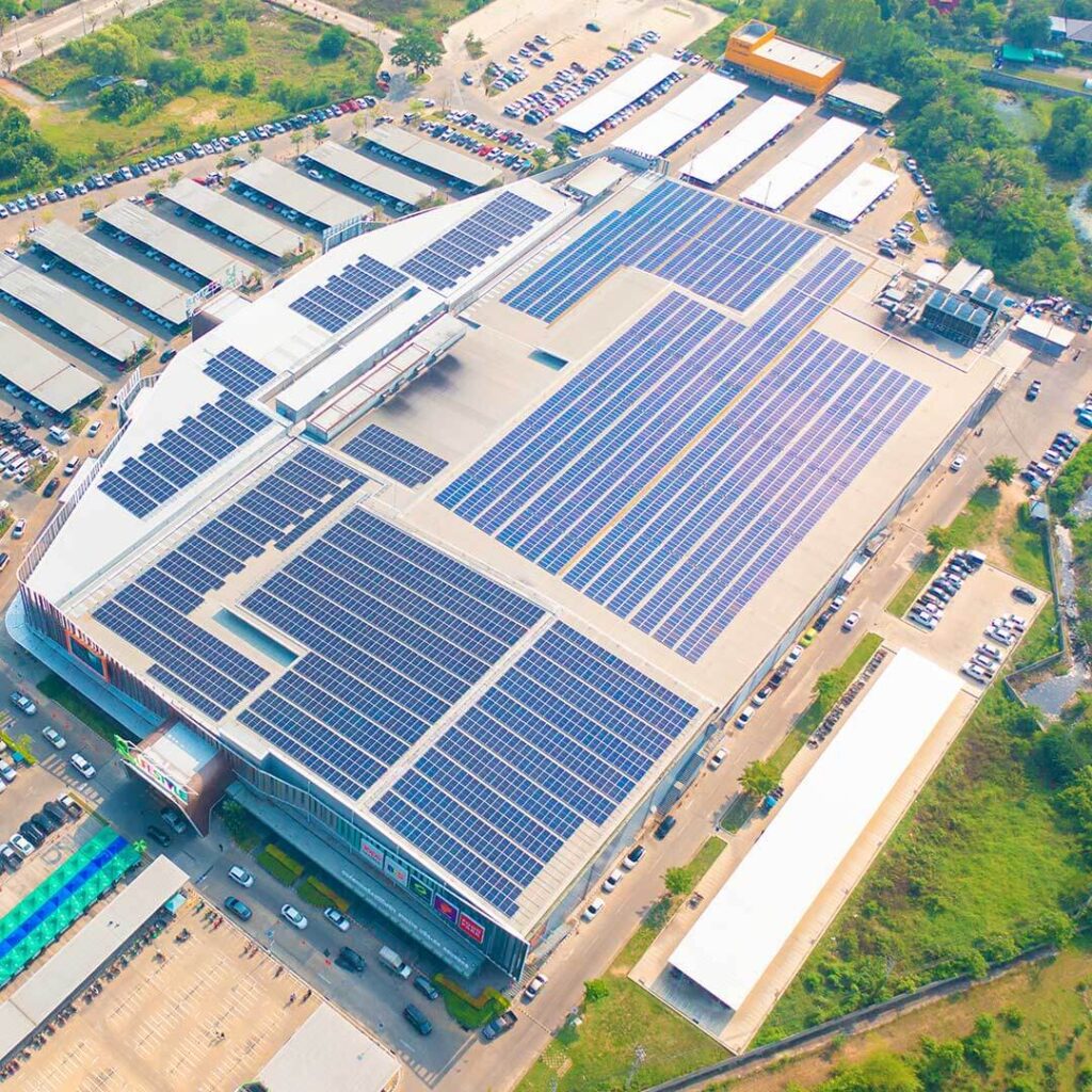 Aerial view of a large commercial facility with extensive solar panels covering the rooftop, surrounded by parking lots and green spaces. The solar installation highlights the site's focus on renewable energy and sustainability.