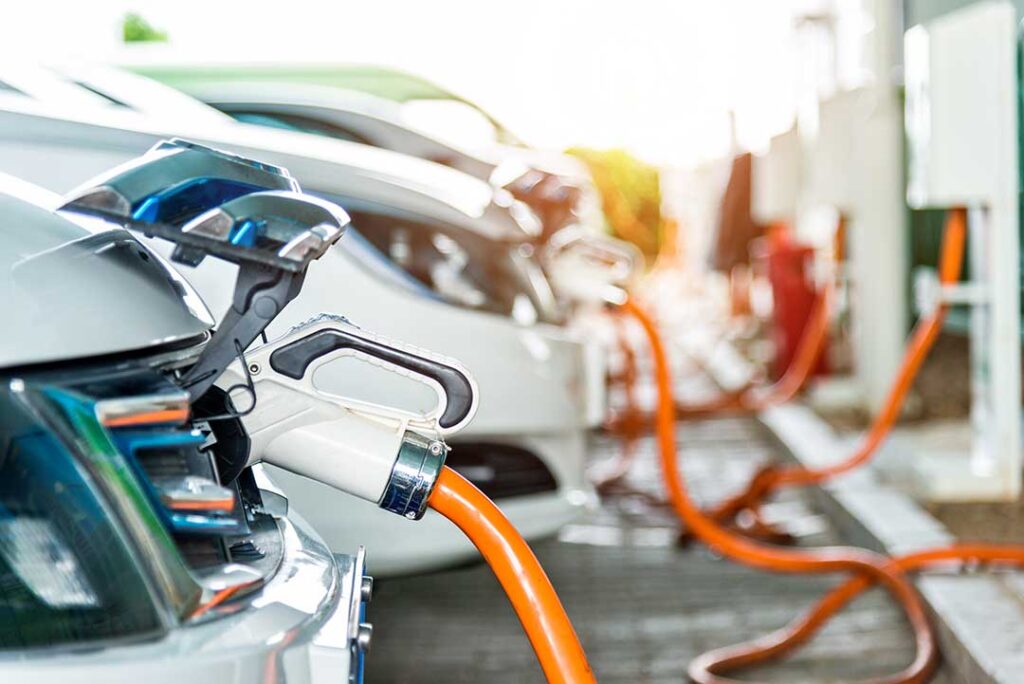 Close-up of an electric vehicle plugged into a charging station, highlighting the bright orange cable and modern connector design, with a row of additional EVs charging in the background under natural sunlight.